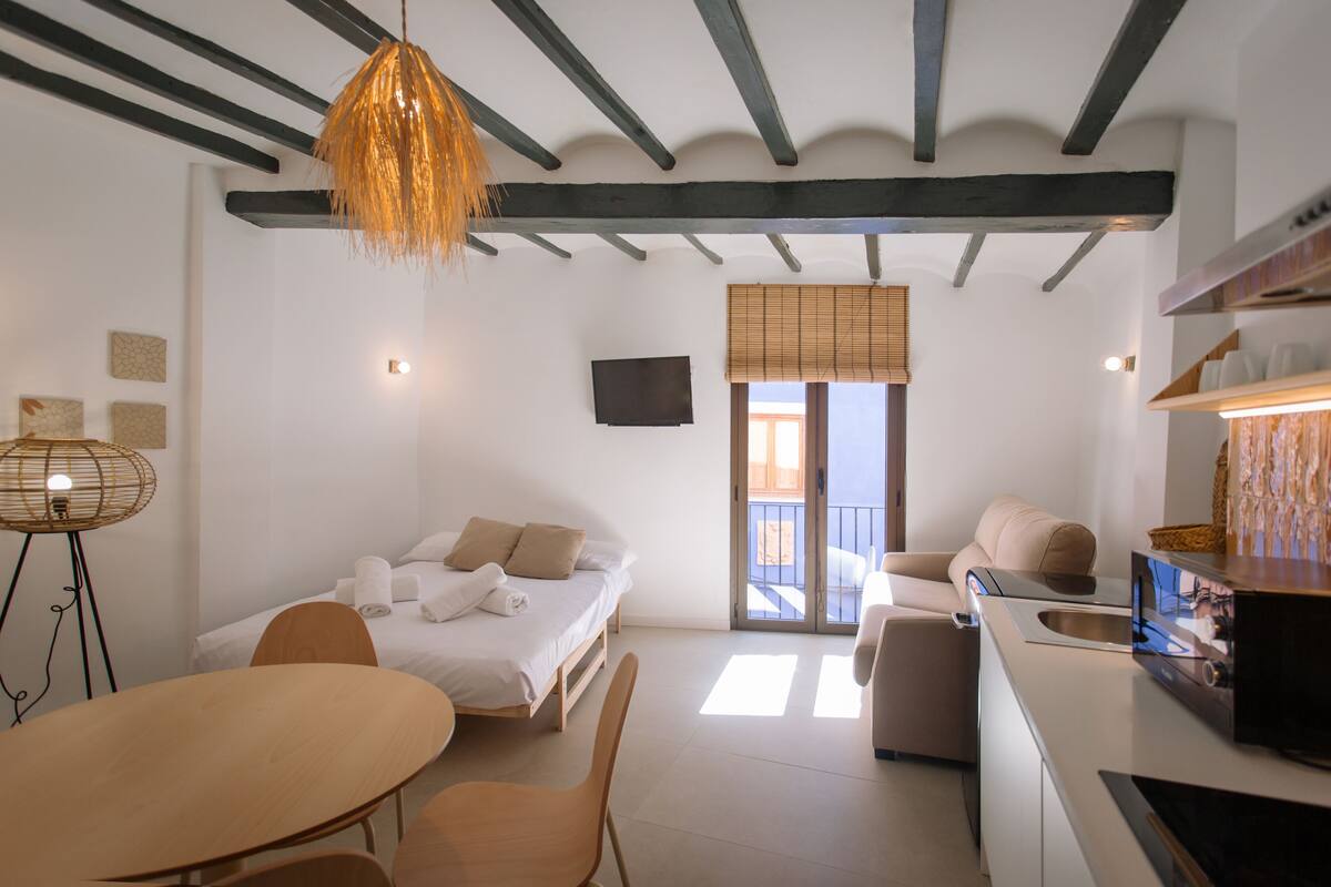 A modern hotel room with a bed, a dining table, a sofa, a kitchen area, and a balcony with a view.