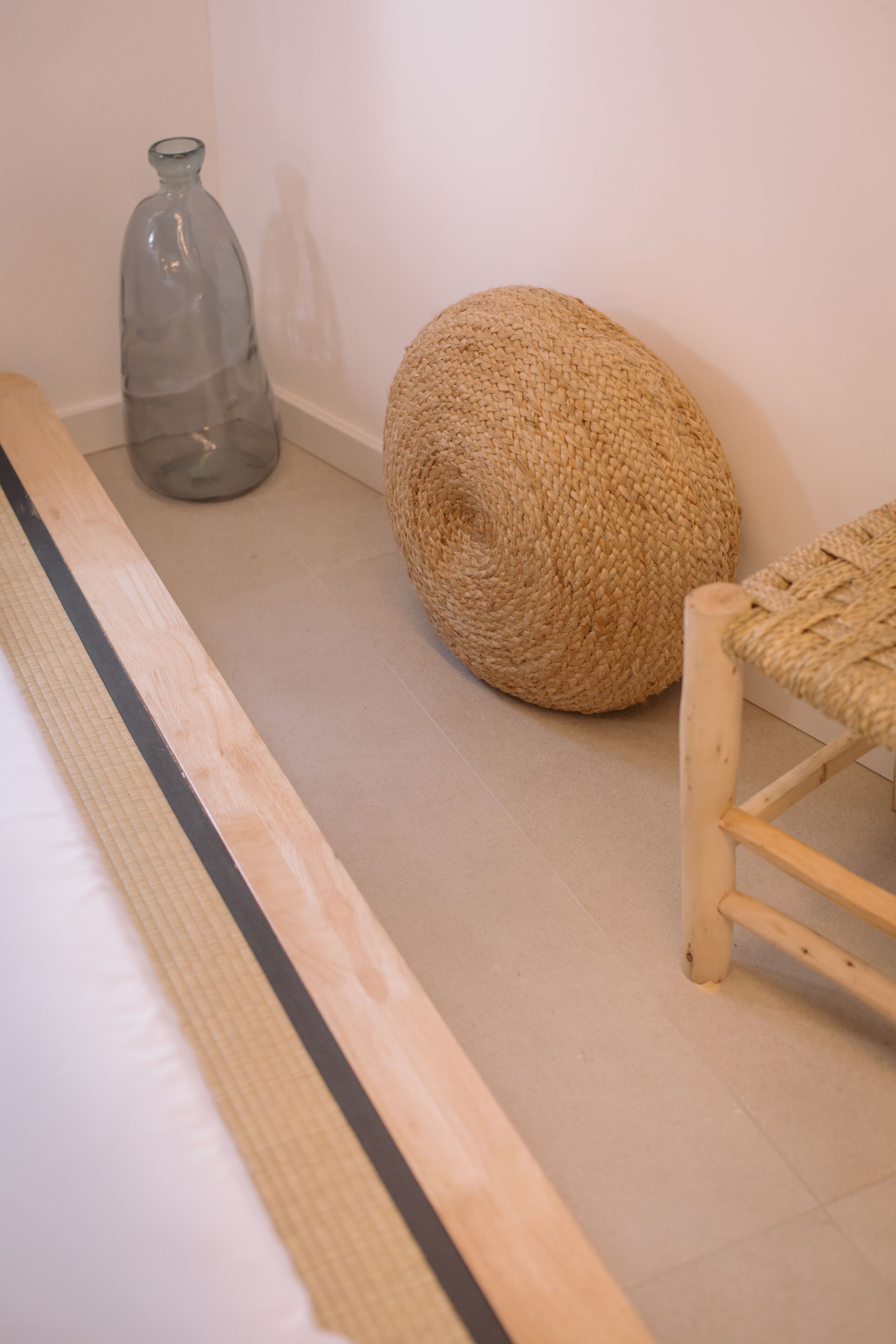 Une pièce avec un plancher en bois, un pouf en fibres tressées, une bouteille en verre et une chaise en bois.