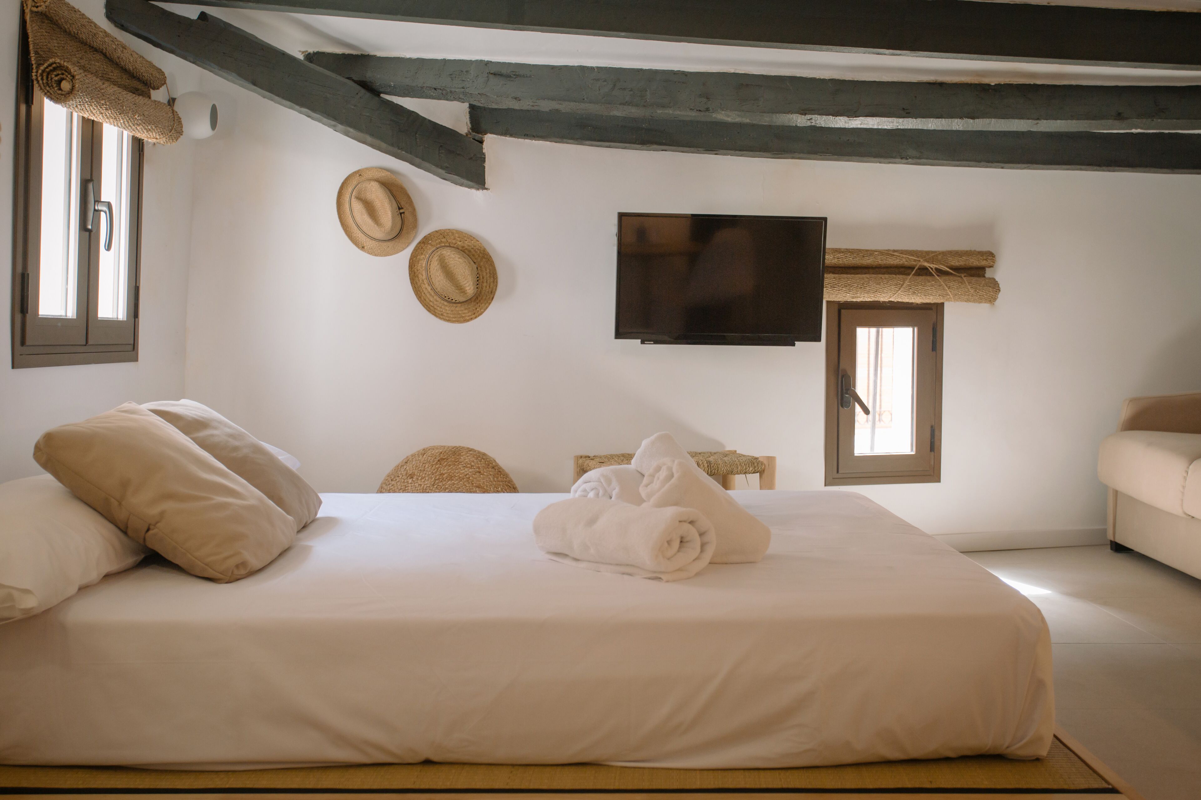 A neatly made bed with pillows and a rolled towel, a television on the wall, and two windows with curtains.