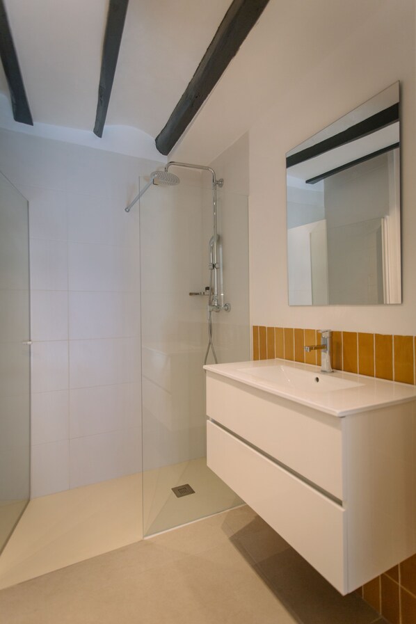 Un baño moderno con ducha envuelta en vidrio, tocador blanco con espejo y suelo de baldosas.