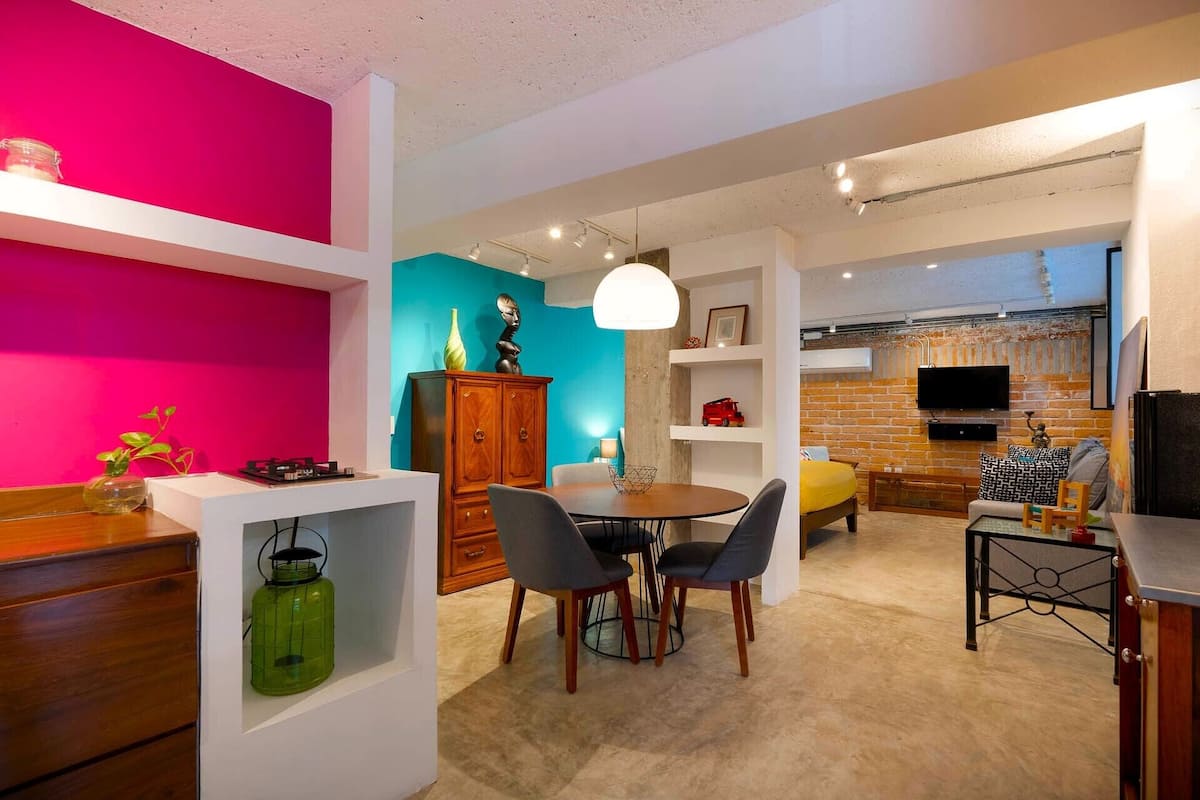 A modern living room with a dining area, a kitchen, and a TV mounted on a brick wall.