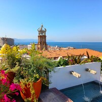 Una piscina en la azotea con vistas despejadas a una ciudad costera, que incluye una torre del reloj destacada y edificios con tejados de teja.