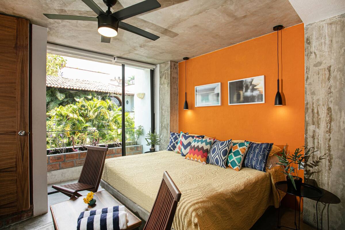 A bedroom with a bed, a ceiling fan, a window with a view of greenery, and a small table with a chair.