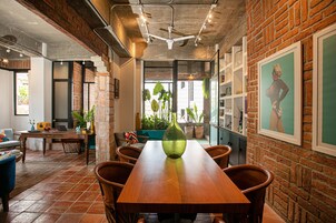 Un comedor con una mesa de madera, sillas y un florero verde. Se ven plantas en macetas y una pared de ladrillo al fondo.