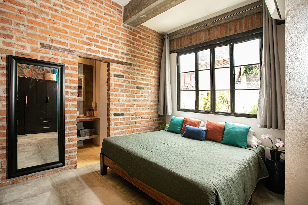A bedroom with a brick wall, a large window, a bed with multiple pillows, and a mirror.