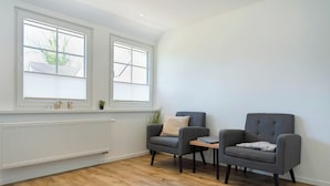 A room with two grey armchairs, a small table, a radiator, and large windows with blinds. - The approx. 45 m² large and very chic 2-room apartment for 1 to 2 persons is located on the Klosterhof Aue in Bad Zwischenahn. (Bad Zwischenahn)