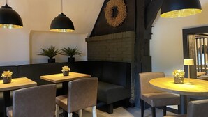 A cozy restaurant interior with wooden tables and chairs, black pendant lights, a brick fireplace, and a decorative wreath on the wall. - The approx. 45 m² large and very chic 2-room apartment for 1 to 2 persons is located on the Klosterhof Aue in Bad Zwischenahn. (Bad Zwischenahn)