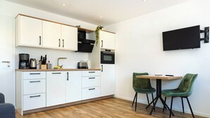 A modern kitchen with white cabinets, a coffee maker, and a microwave. There is a dining area with two green chairs and a table. - The approx. 45 m² large and very chic 2-room apartment for 1 to 2 persons is located on the Klosterhof Aue in Bad Zwischenahn. (Bad Zwischenahn)