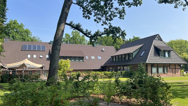 A two-story house with a sloped roof, a covered patio area, and a well-maintained garden with young trees and a paved walkway. - The approx. 45 m² large and very chic 2-room apartment for 1 to 2 persons is located on the Klosterhof Aue in Bad Zwischenahn. (Bad Zwischenahn)