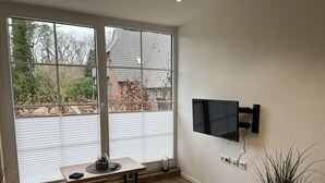 Una habitación con ventanas amplias, un televisor de pantalla plana en la pared, una mesa de madera con un florero y un control remoto, y una planta en maceta.