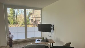 Una sala de estar con un ventanal grande, un televisor montado, una mesita con un control remoto y un sofá.
