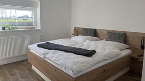 Un dormitorio moderno con estructura de cama de madera, un ventanal con persianas, un radiador y una mesita de noche con lámpara.