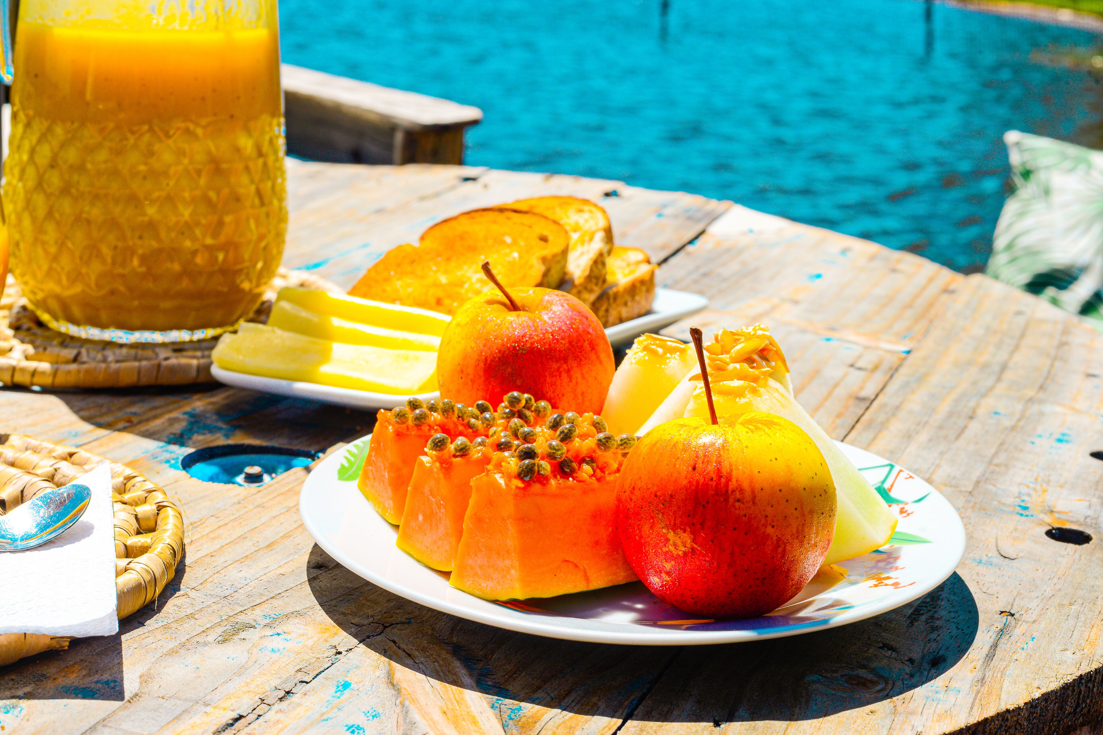 Desayuno con una fuente de fruta, tostadas y un vaso de zumo de naranja junto a una piscina.