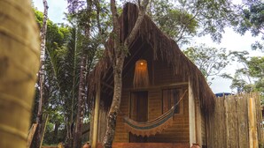 Una cabaña de madera con techo de paja, rodeada de árboles y una hamaca.