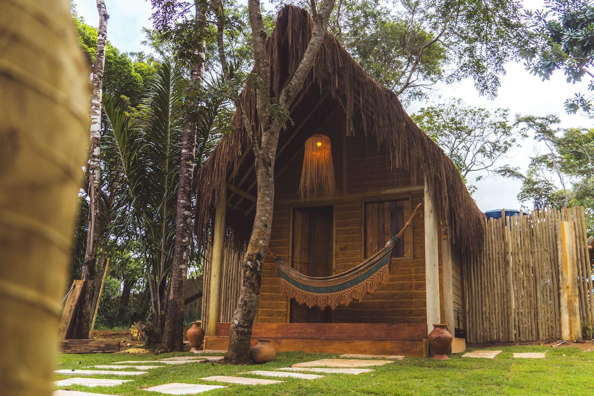 Una cabaña de madera con techo de paja, rodeada de árboles y una hamaca.