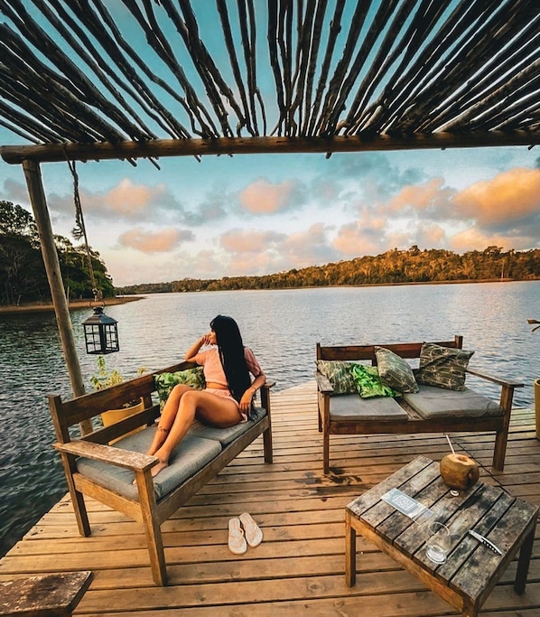 Una terraza de madera con un banco y una mesa, con vista a un cuerpo de agua tranquilo y una orilla boscosa.