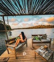 A wooden deck with a bench and a table, overlooking a calm body of water and a forested shoreline.