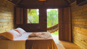 Un dormitorio de cabaña de madera con una cama, una hamaca y una ventana con vistas a follaje.