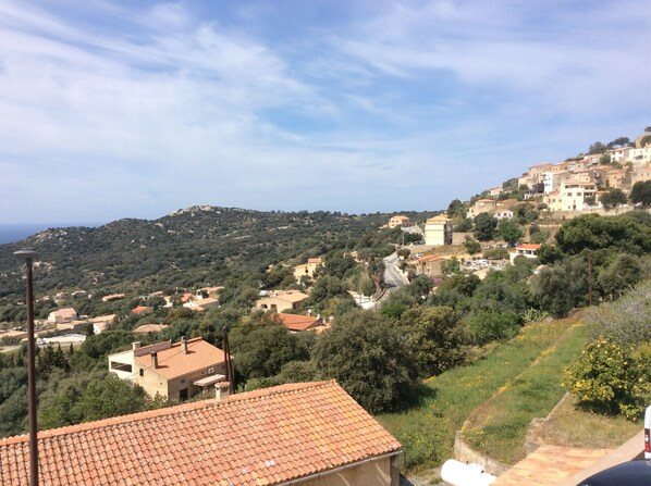 A hillside village with terracotta-roofed buildings, a clear sky, and a distant view of the sea.