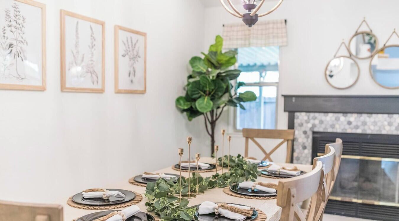 Et spiseområde med et bord dækket til fire personer, omgivet af stole, vægmalerier med botaniske motiver og en stor plante i midten.