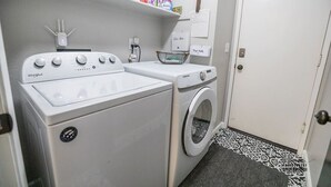 Un área de lavandería con lavadora Whirlpool y secadora Samsung, un estante con productos de limpieza y una puerta con un tapete decorativo.