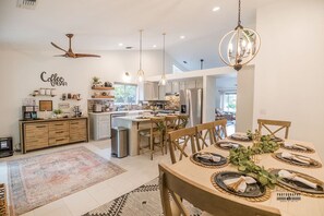 A modern kitchen with a central island, a dining area with a wooden table set for four, and a coffee bar area. - Hanland House | Home Away From Home (Redding)