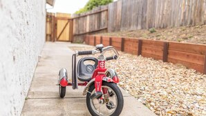 Une moto rouge avec un side-car est garée sur un chemin de béton, à côté d’une clôture en bois.