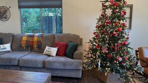 Una sala de estar con un sofá gris, un árbol de Navidad y una ventana con vistas a la vegetación.