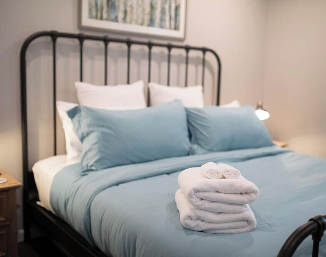 A neatly made bed with a blue bedspread and white pillows, a stack of folded towels, and a metal headboard.