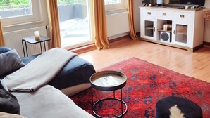 A modern living room with a sofa, ottoman, and a red patterned rug. There is a television on a white cabinet, and large windows with curtains.