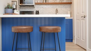 Una cocina con armarios azules, una encimera blanca, dos taburetes marrones, un microondas y una cafetera.