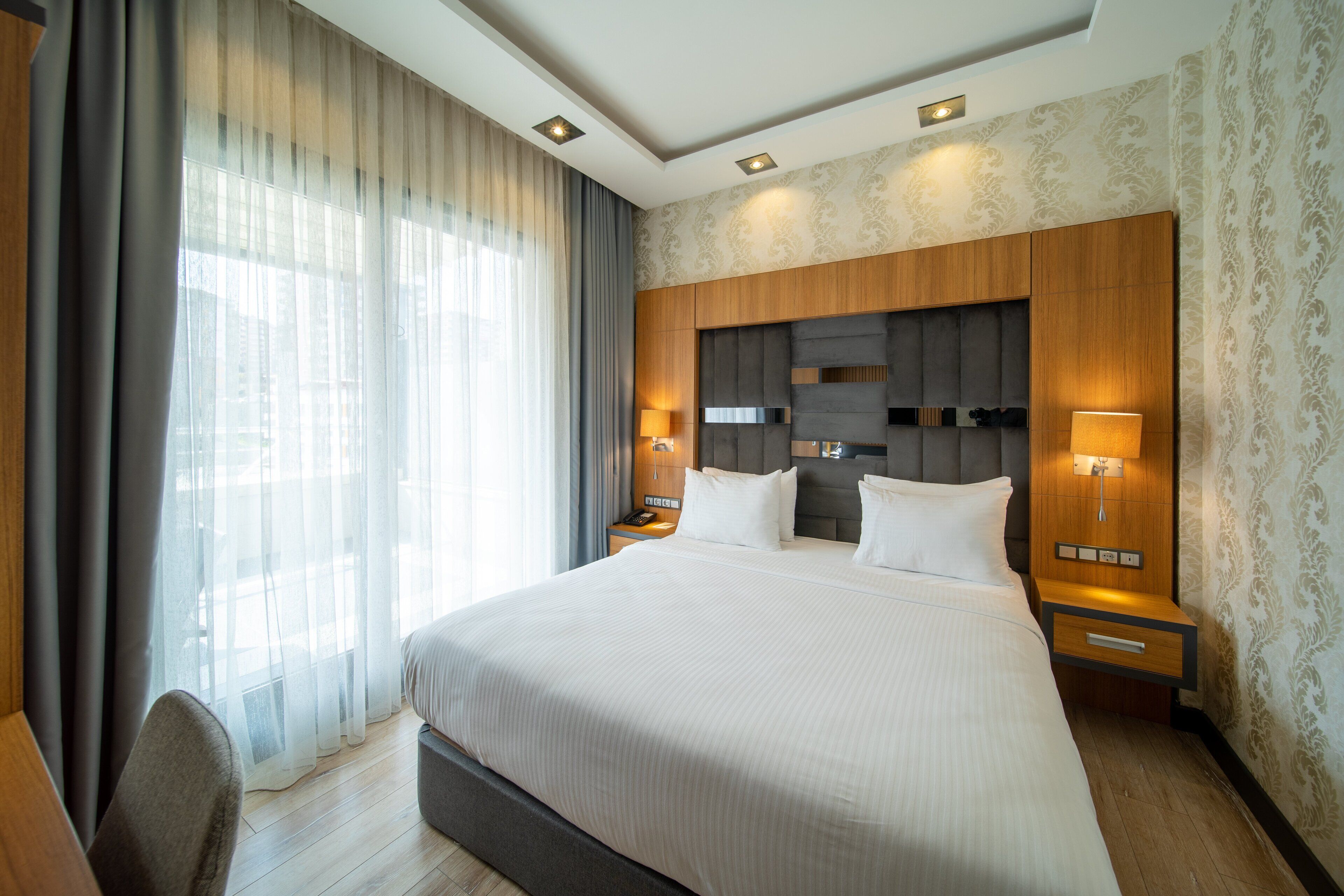 A modern hotel room with a large bed, wooden headboard, two bedside tables with lamps, a chair, and a large window with sheer curtains.