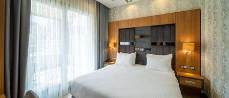 A modern hotel room with a large bed, wooden headboard, two bedside tables with lamps, a chair, and a large window with sheer curtains.