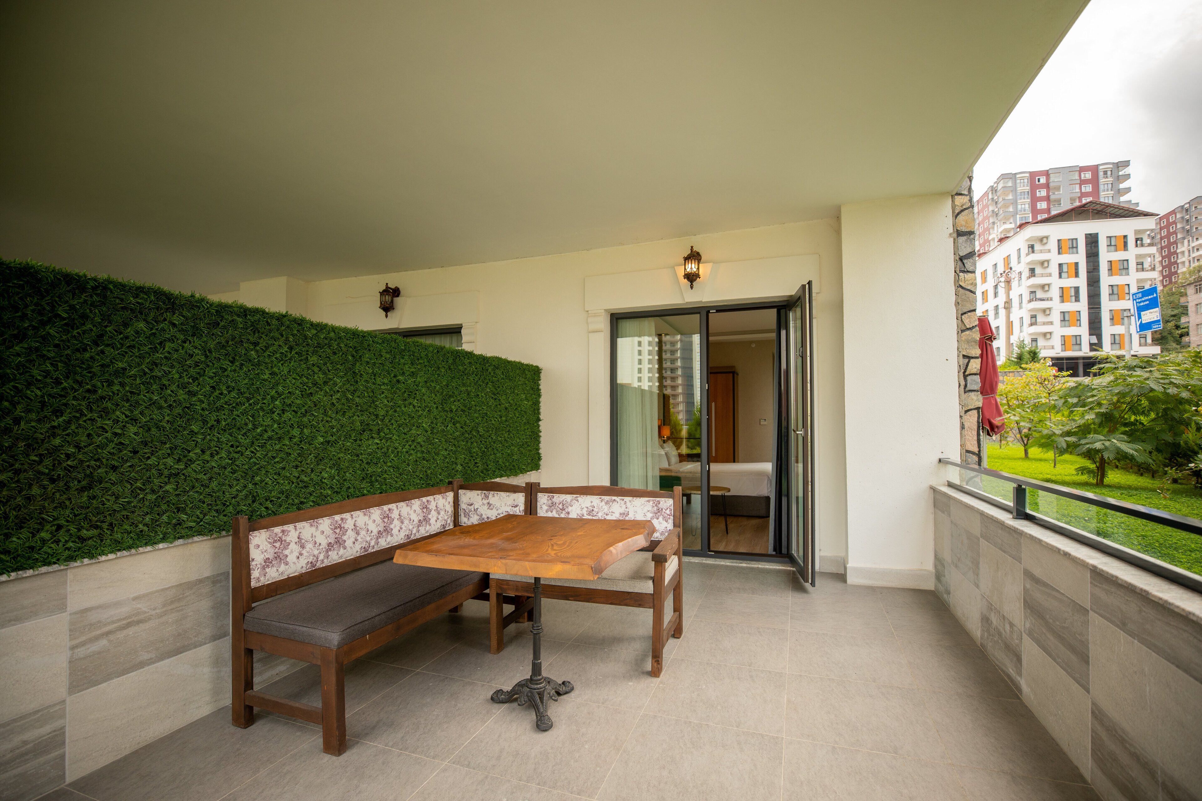 A balcony with a wooden bench and table, a green hedge, and a view of a building in the background.