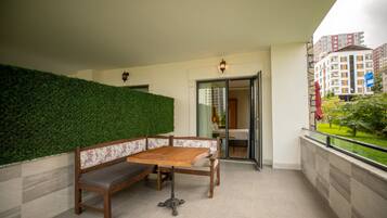 A balcony with a wooden bench and table, a green hedge, and a view of a building in the background.