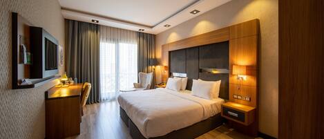 A hotel room with a large bed, a desk with a lamp, a TV, and a nightstand with a lamp.