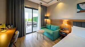 A hotel room with a wooden desk, a green sofa, a small round table, a bed, a nightstand with a lamp, and a view of a balcony with outdoor furniture. - Holiday Inn Trabzon East by IHG (Yomra)