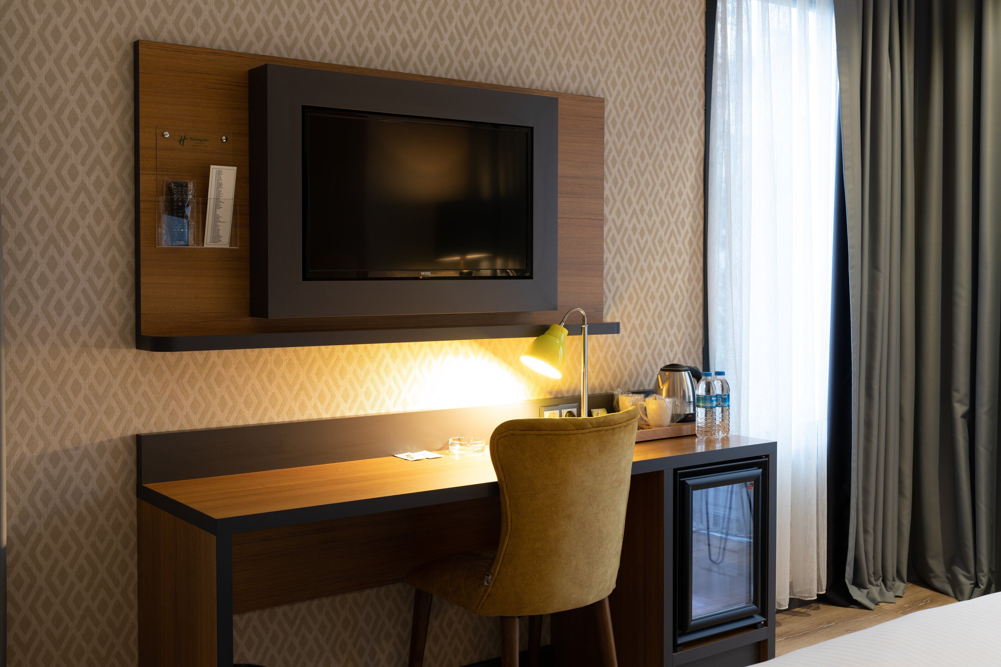 A hotel room with a wooden desk, a television mounted on the wall, a chair, a lamp, and a bed.