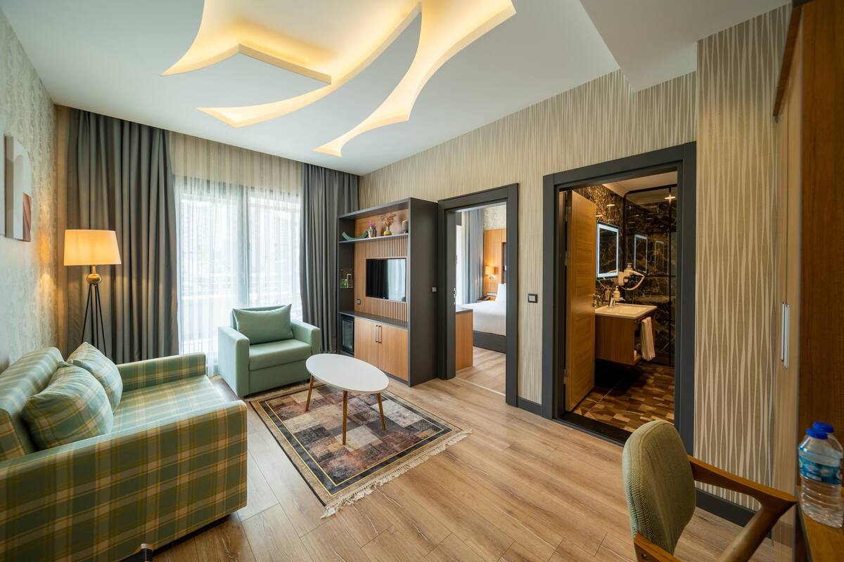 Une chambre d’hôtel moderne avec un canapé à carreaux verts, une petite table ronde et un fauteuil vert. La chambre est dotée d’un parquet, d’un tapis à motifs et d’un grand miroir.