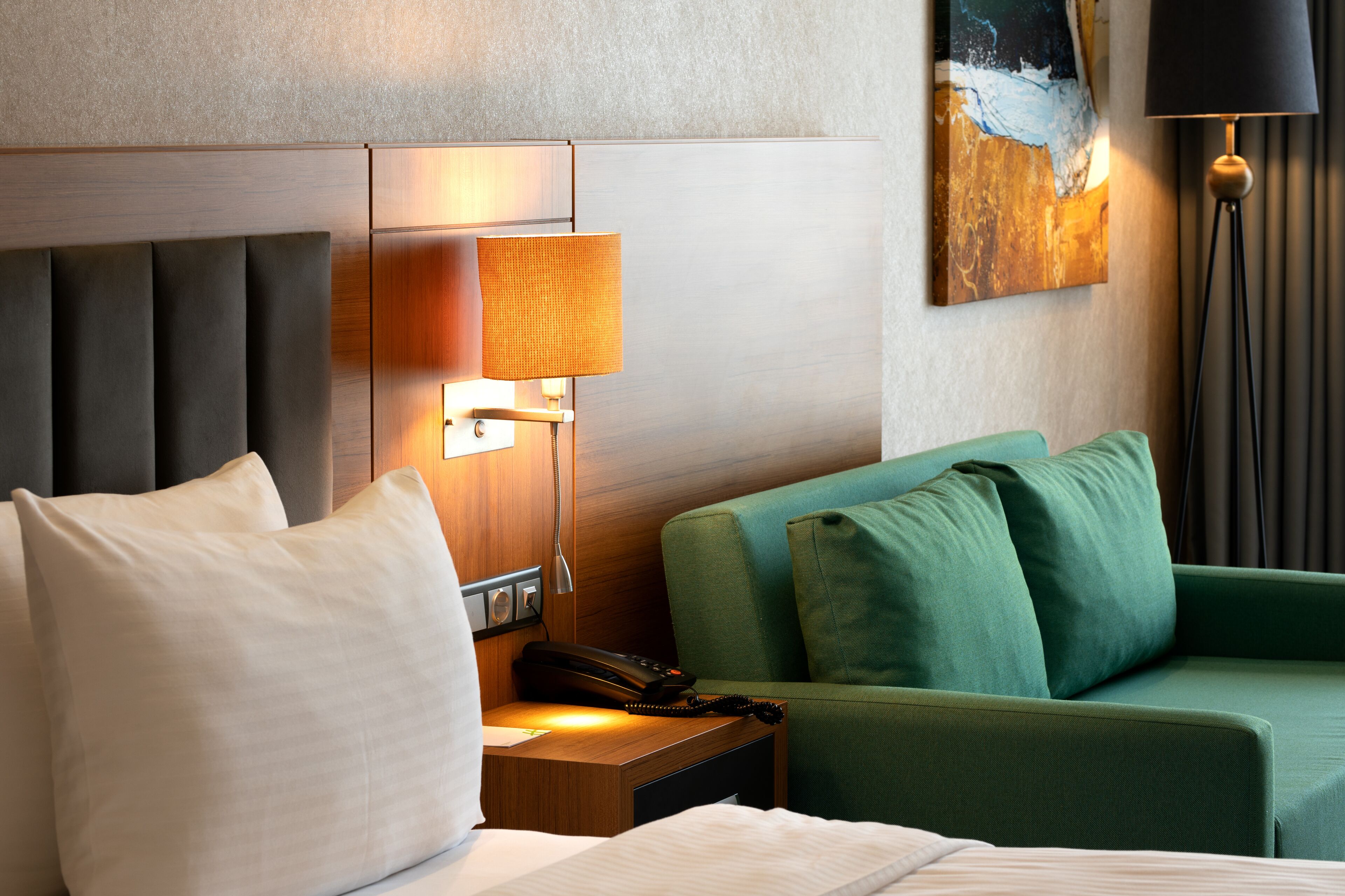 Habitación de hotel con cama, sofá verde, mesita de noche con lámpara y un cuadro en la pared.