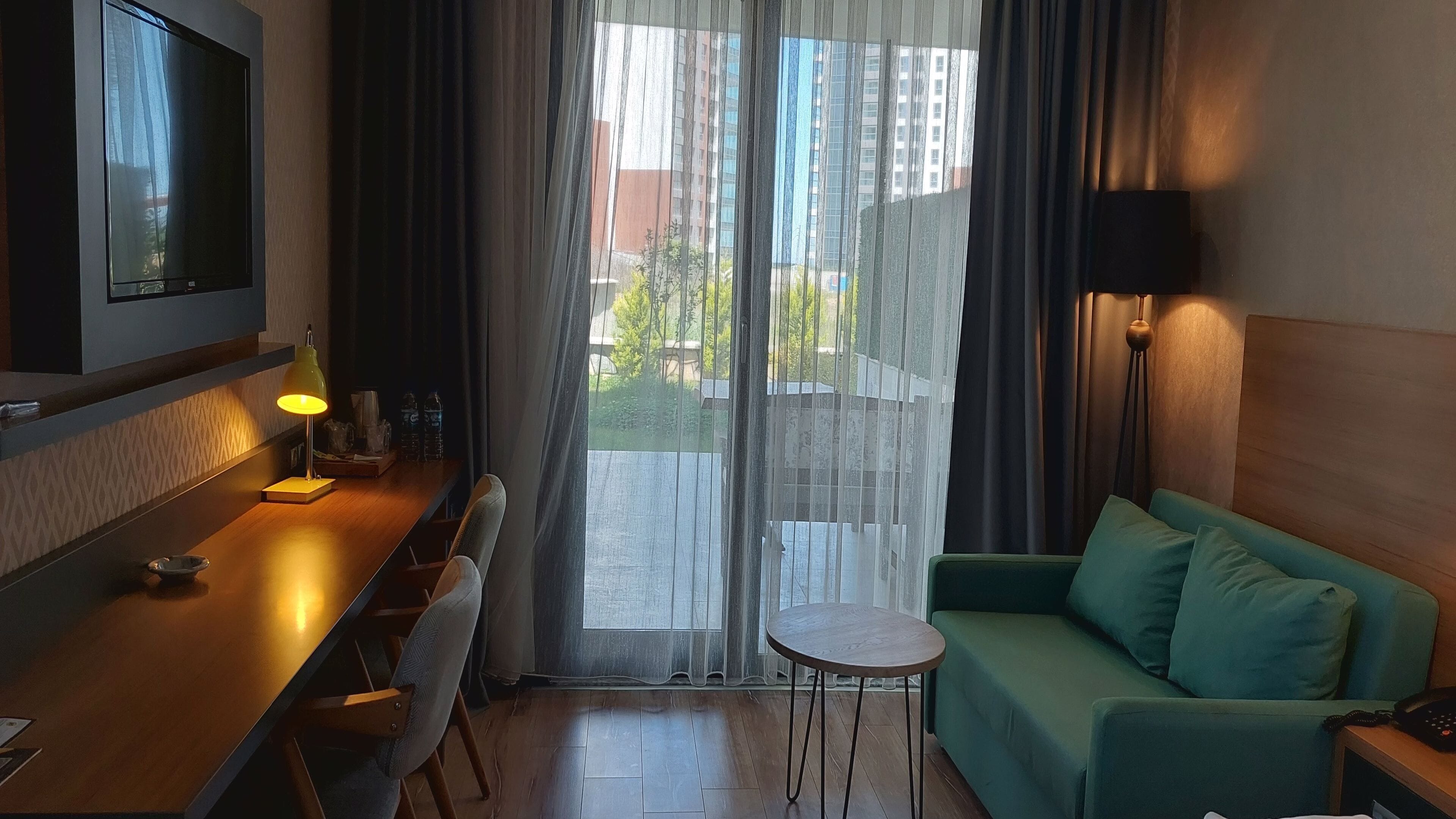 A hotel room with a wooden desk, a green sofa, a small round table, a TV mounted on the wall, and a view of the outside through sheer curtains.