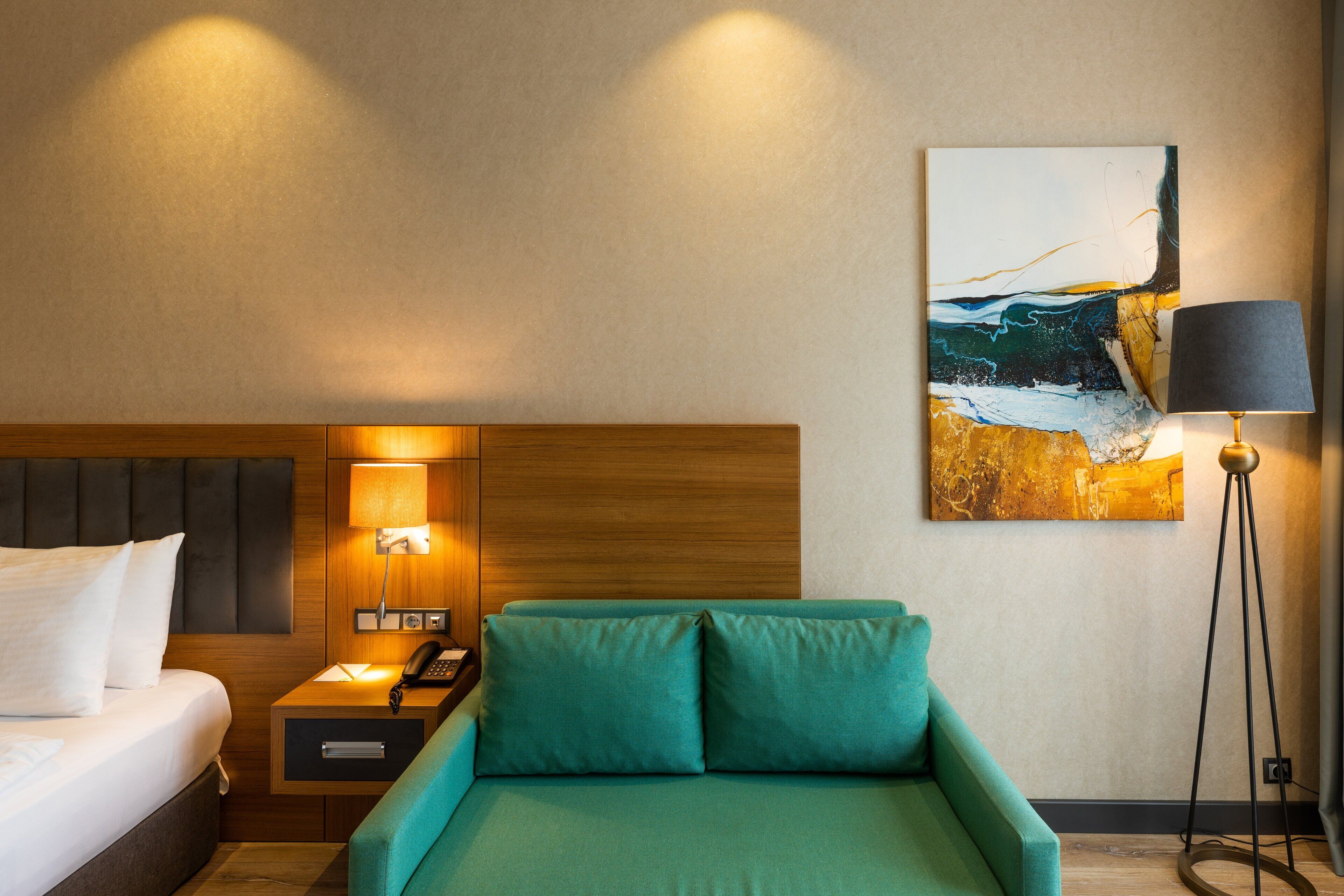 Una habitación de hotel con una cama, un sofá verde, una mesita de noche con lámpara y un cuadro en la pared.