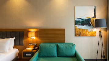 Una habitación de hotel con una cama, un sofá verde, una mesita de noche con lámpara y un cuadro en la pared.