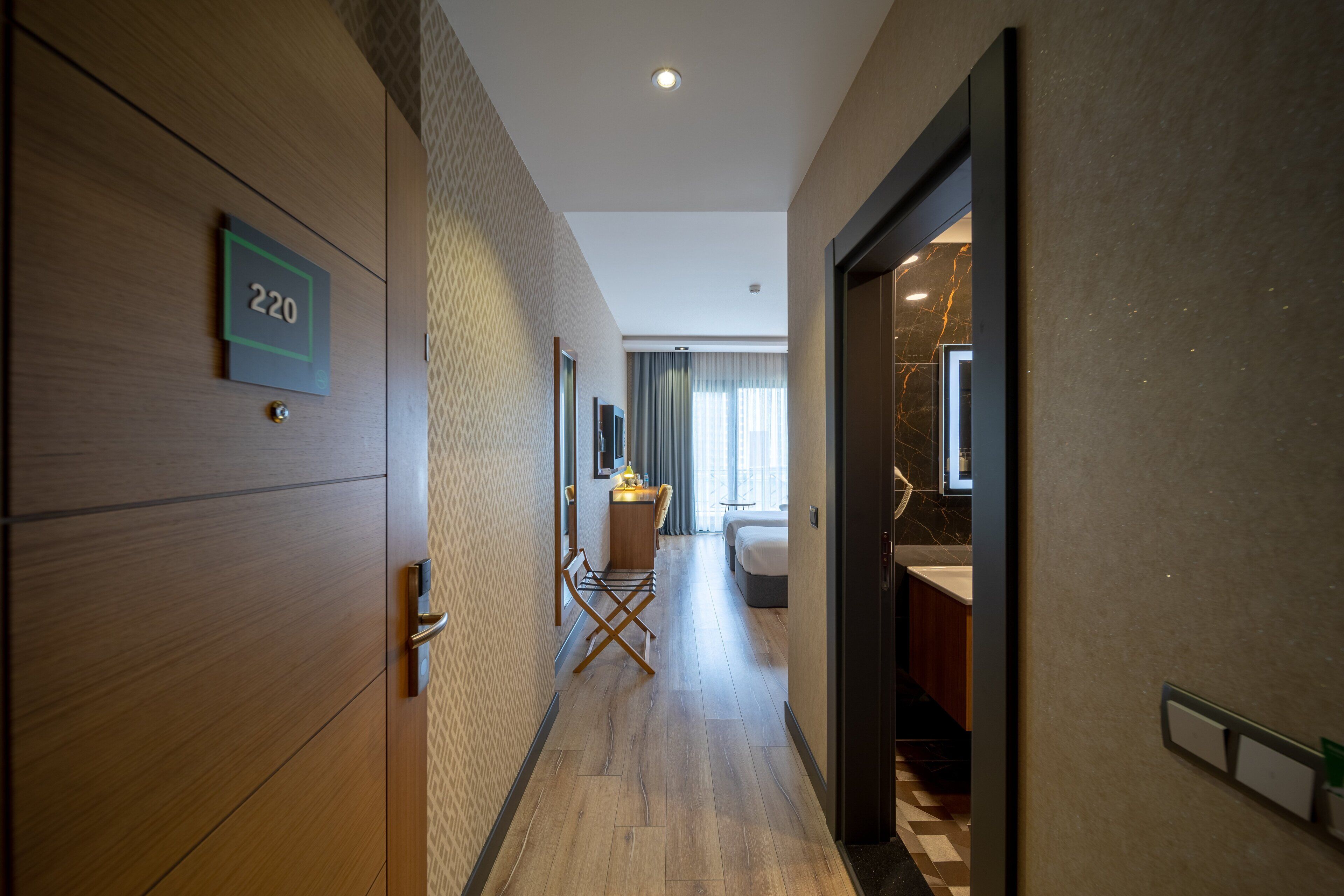 A hotel corridor with a wooden door labeled '220', a seating area with a desk and chair, and a bed in the background.