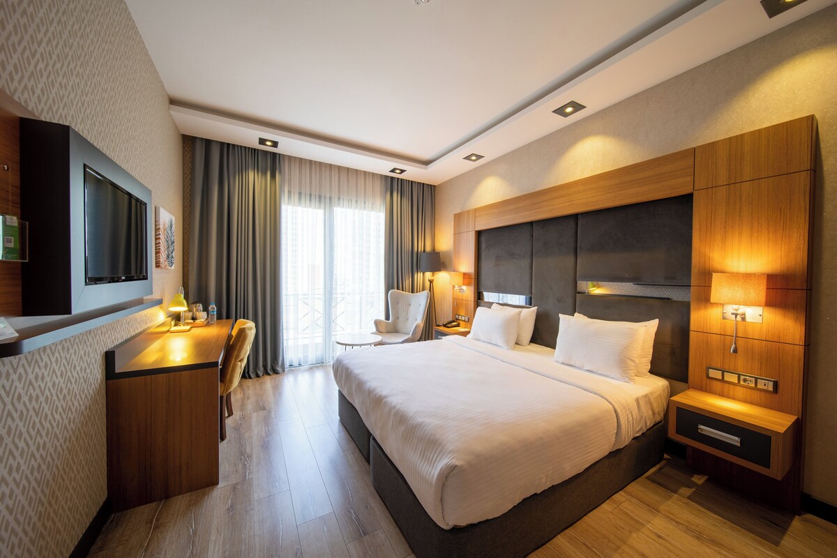 Ein Hotelzimmer mit einem großen Bett, einem Schreibtisch, einem Sessel, einem Fernseher und einem Fenster mit Vorhängen.