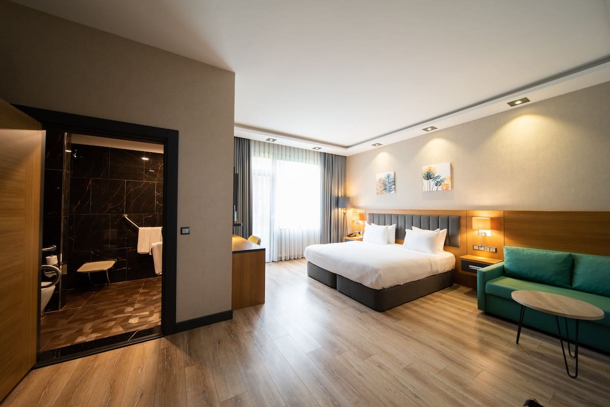 Une chambre d’hôtel moderne avec un grand lit, un canapé vert, une petite table et une salle de bain dotée d’une douche en marbre.