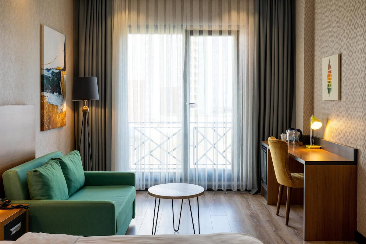 Une chambre d’hôtel avec un canapé vert, une petite table ronde, un bureau avec une chaise et une grande fenêtre avec des rideaux.