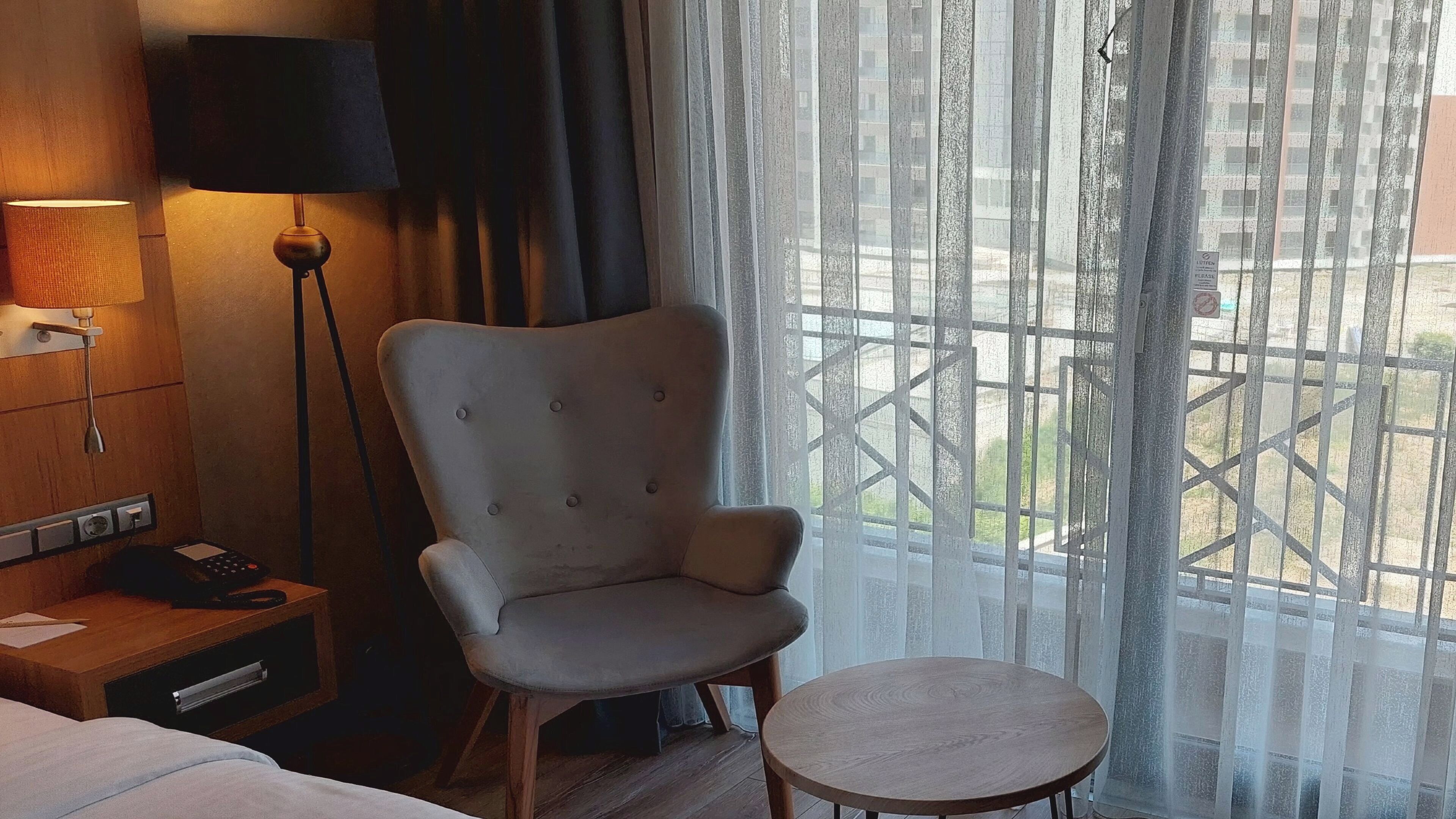 A hotel room with a bed, a chair, a small table, a lamp, a phone, and a window with a view of buildings.