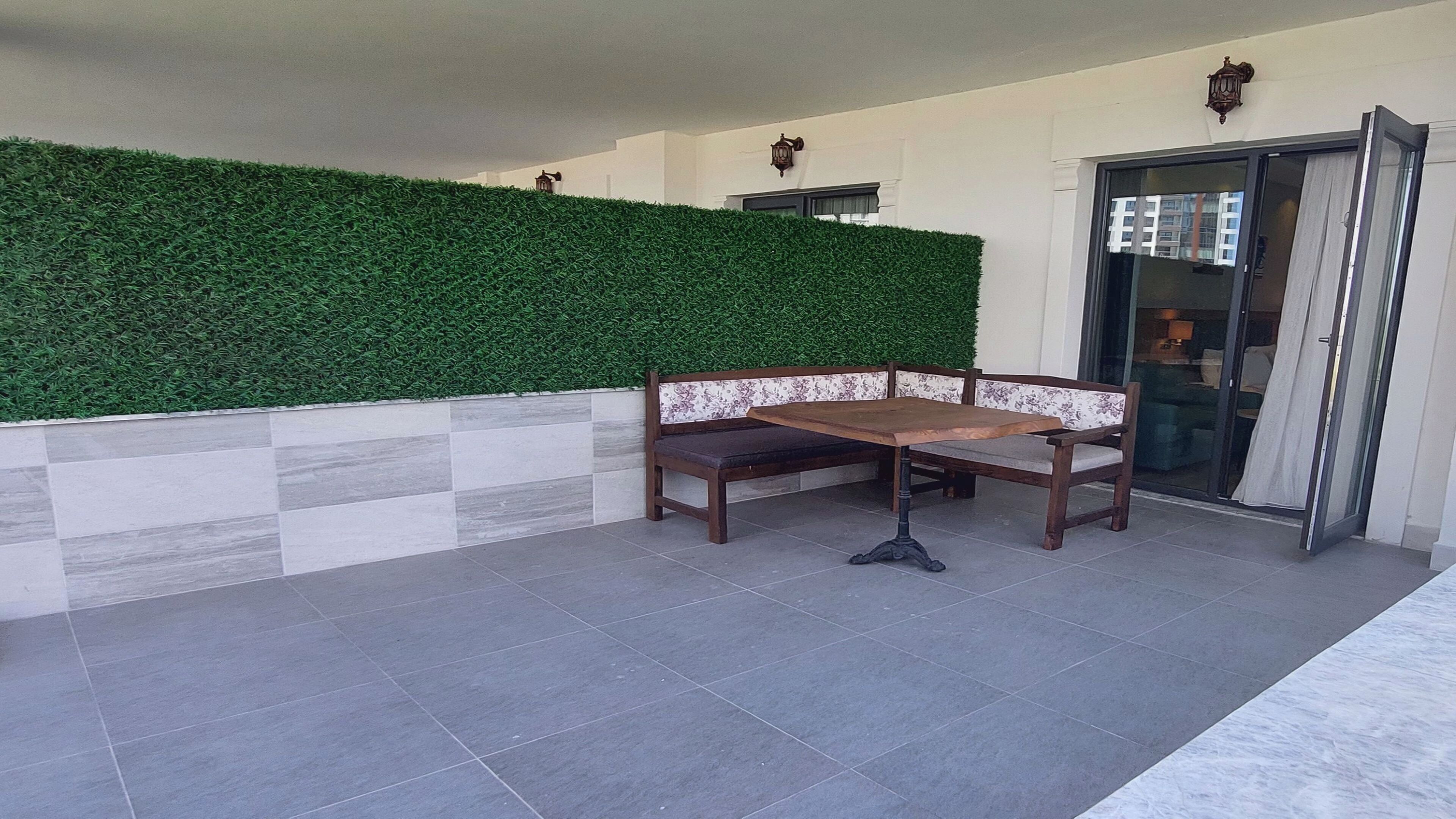 Un espacio de patio con un banco y una mesa, un muro de seto verde y un cerramiento de vidrio corredero que da a un espacio interior.