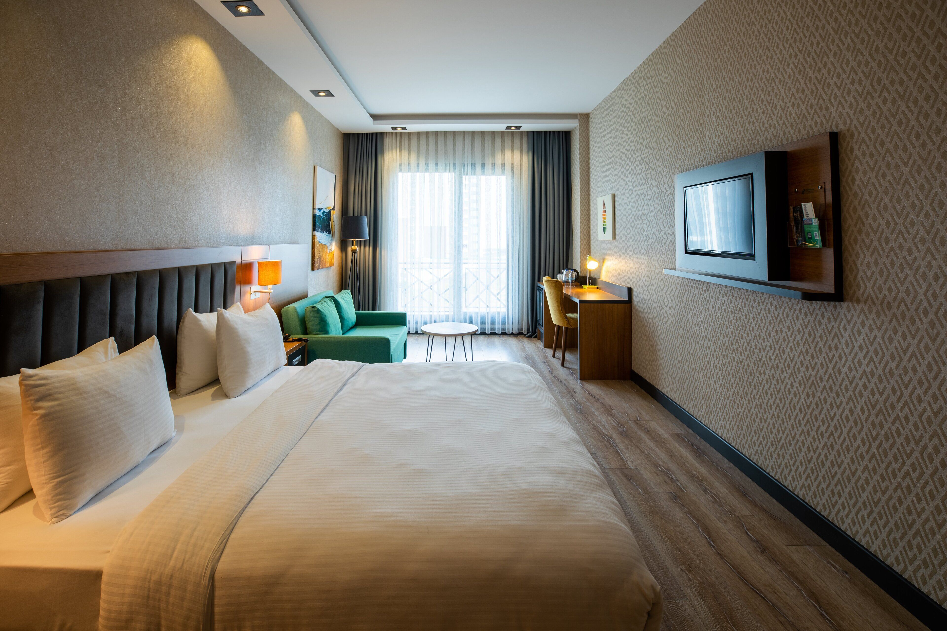 Quarto de hotel com uma cama grande, uma escrivaninha, uma cadeira, um sofá e uma televisão na parede.